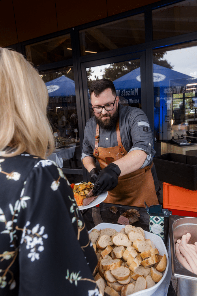 Koch Daniel Parzen mit Lederschürze serviert frisch zubereitete Speisen am Buffet an eine Gästin beim Charity-Golfturnier von Round Table 98 Bayreuth