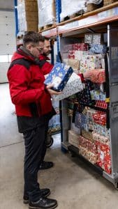 Zwei Helfer in roten Jacken sortieren Weihnachtspäckchen in einen Rollwagen im Lager
